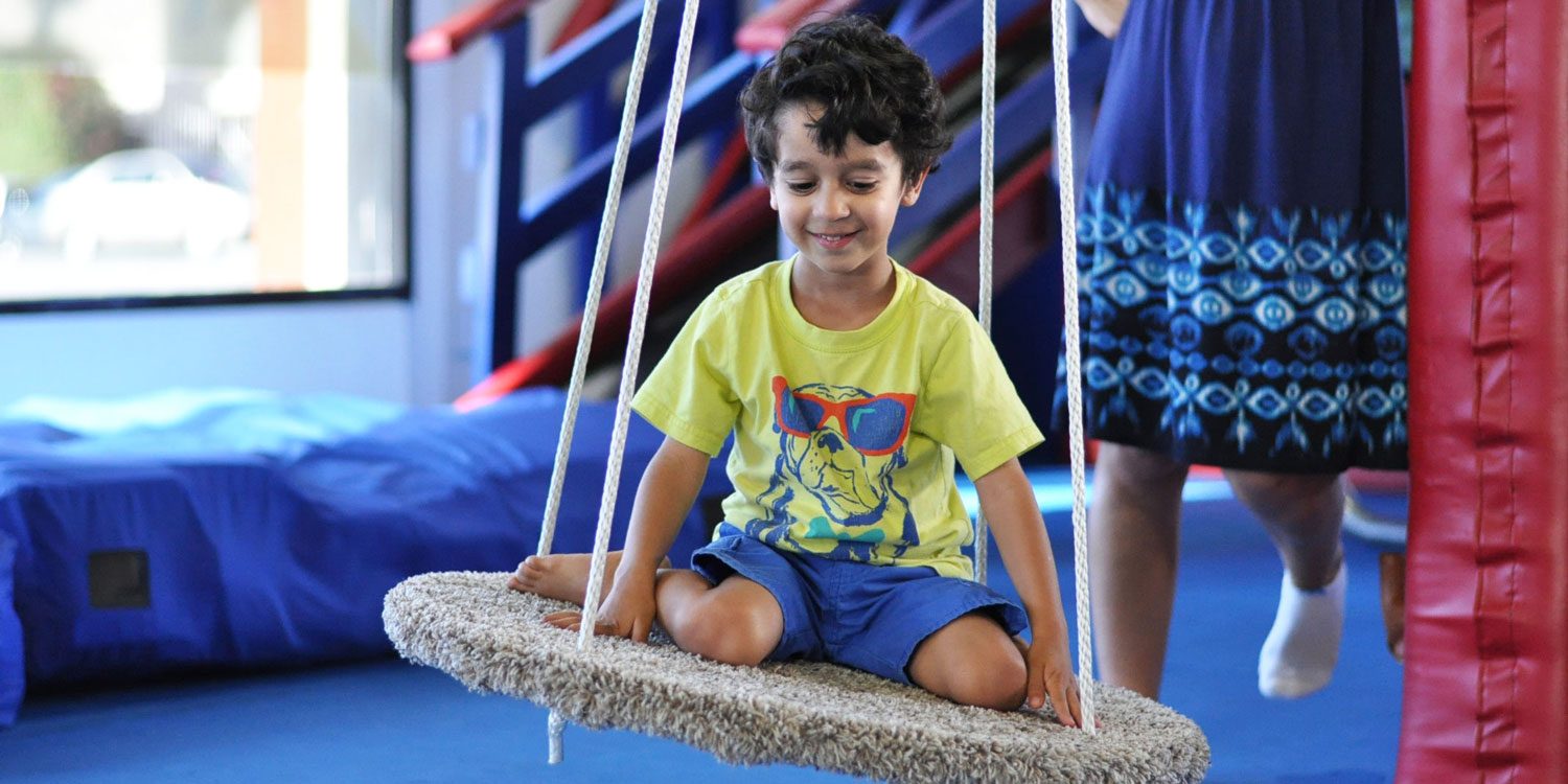 A boy sitting cross-legged on a platform swing in the Joy Therapy sensory gym