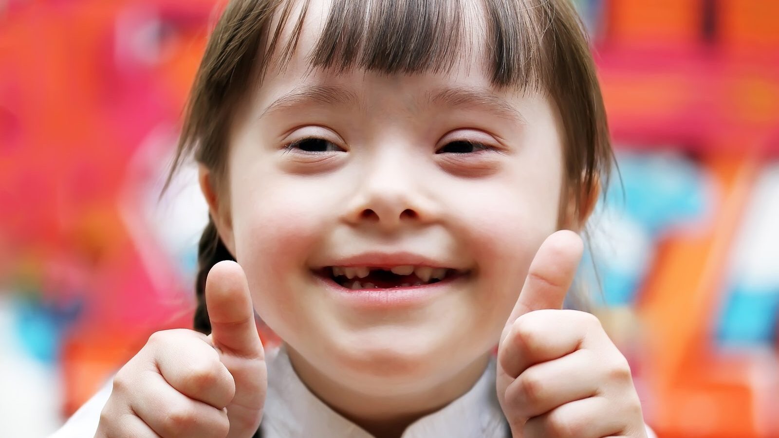 A smiling girl giving two thumbs up in front of a bright, colorful classroom wall