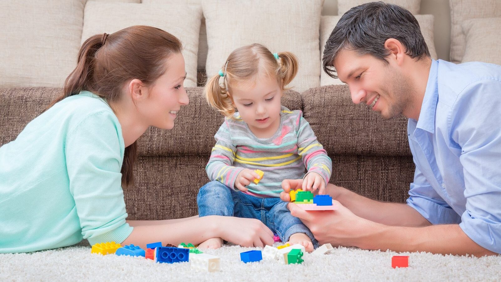 Two parents sitting on the floor with their young daughter, building with colored blocks together