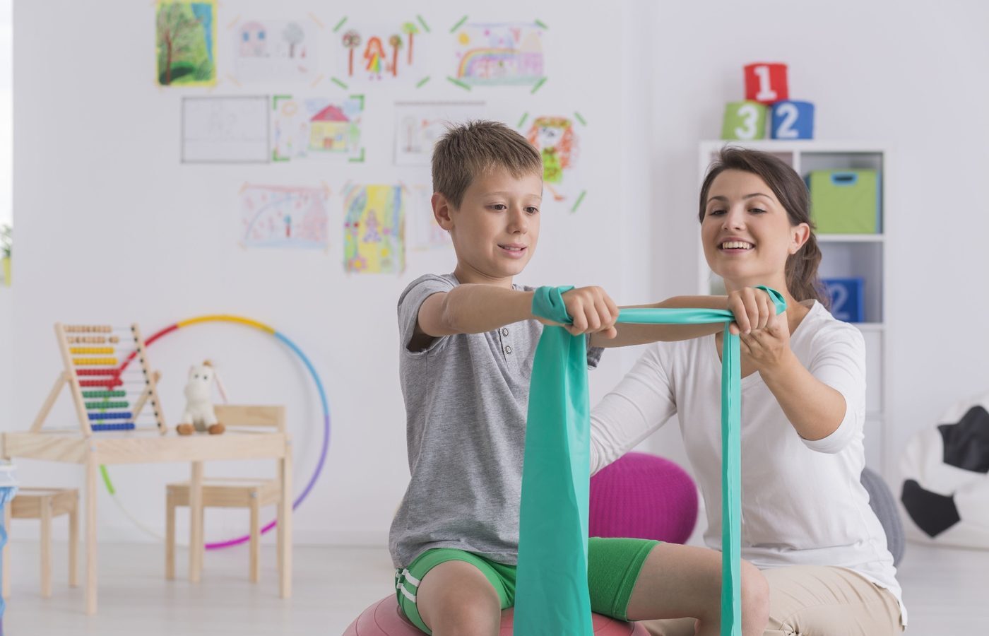 A boy stretching a resistance band with a therapist in a bright, well equipped therapy room