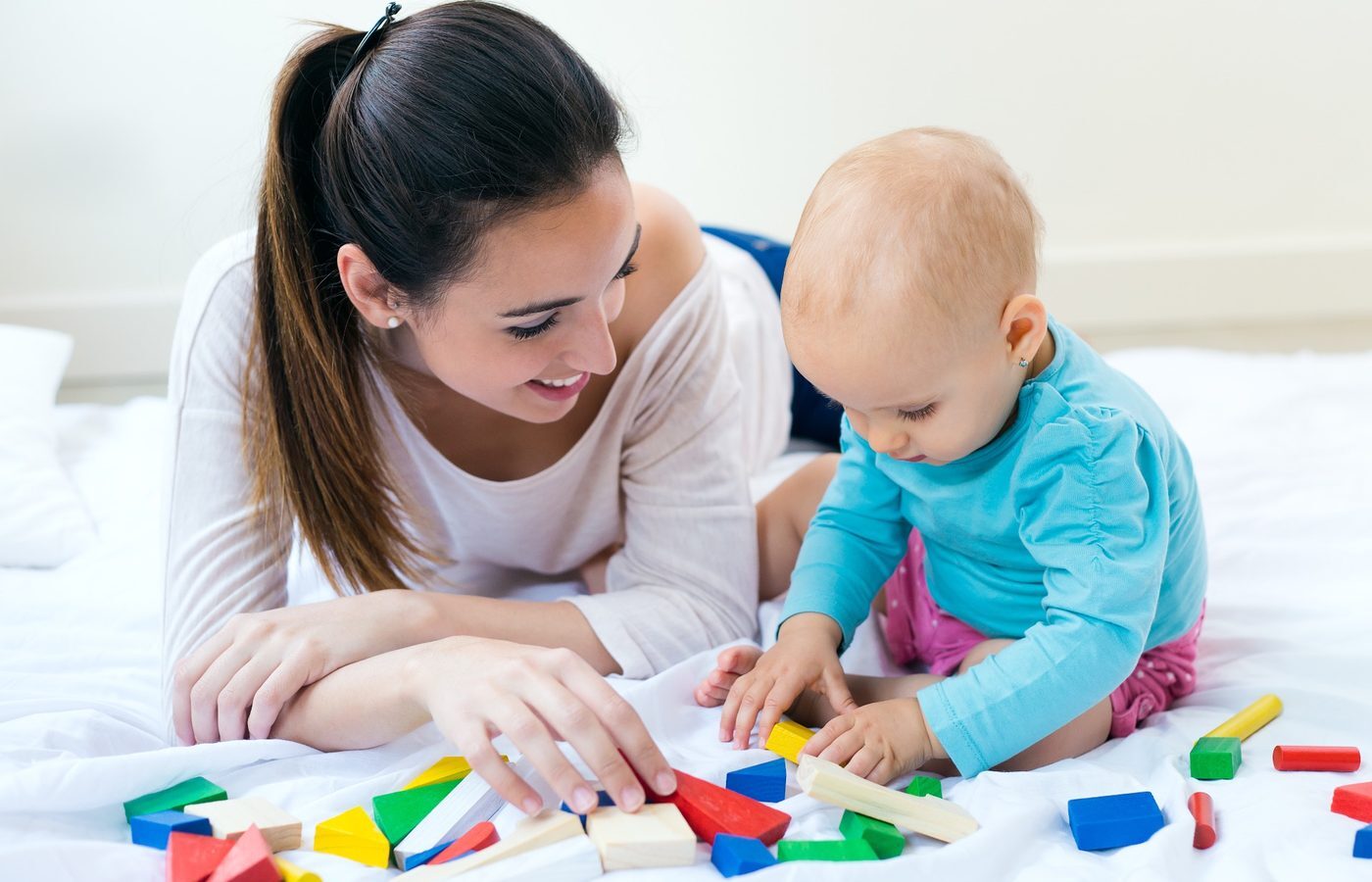 An adult lying on the floor beside a baby, both reaching for colored building blocks