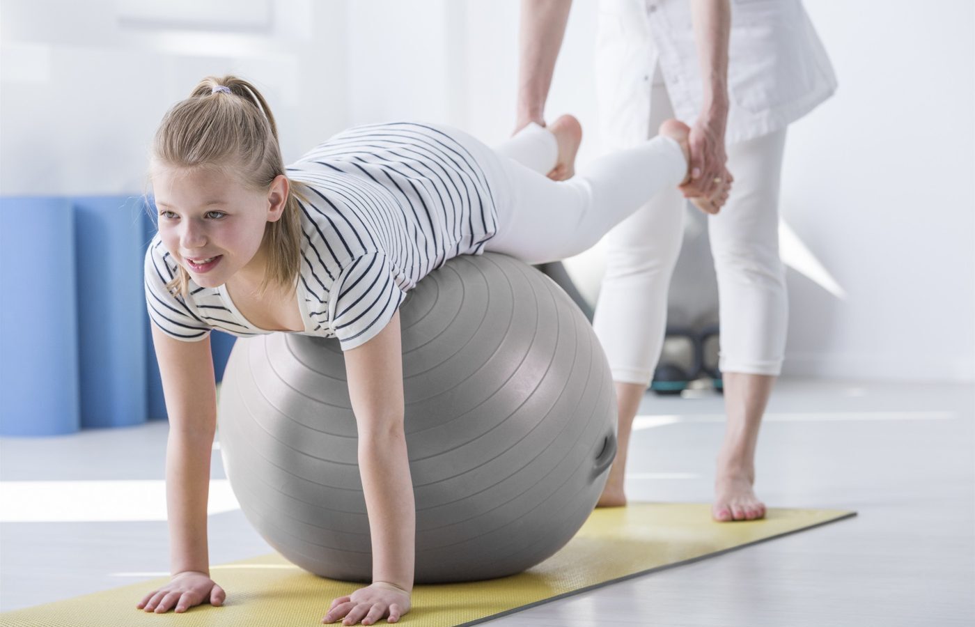 A girl balancing over a large therapy ball while a therapist supports her legs