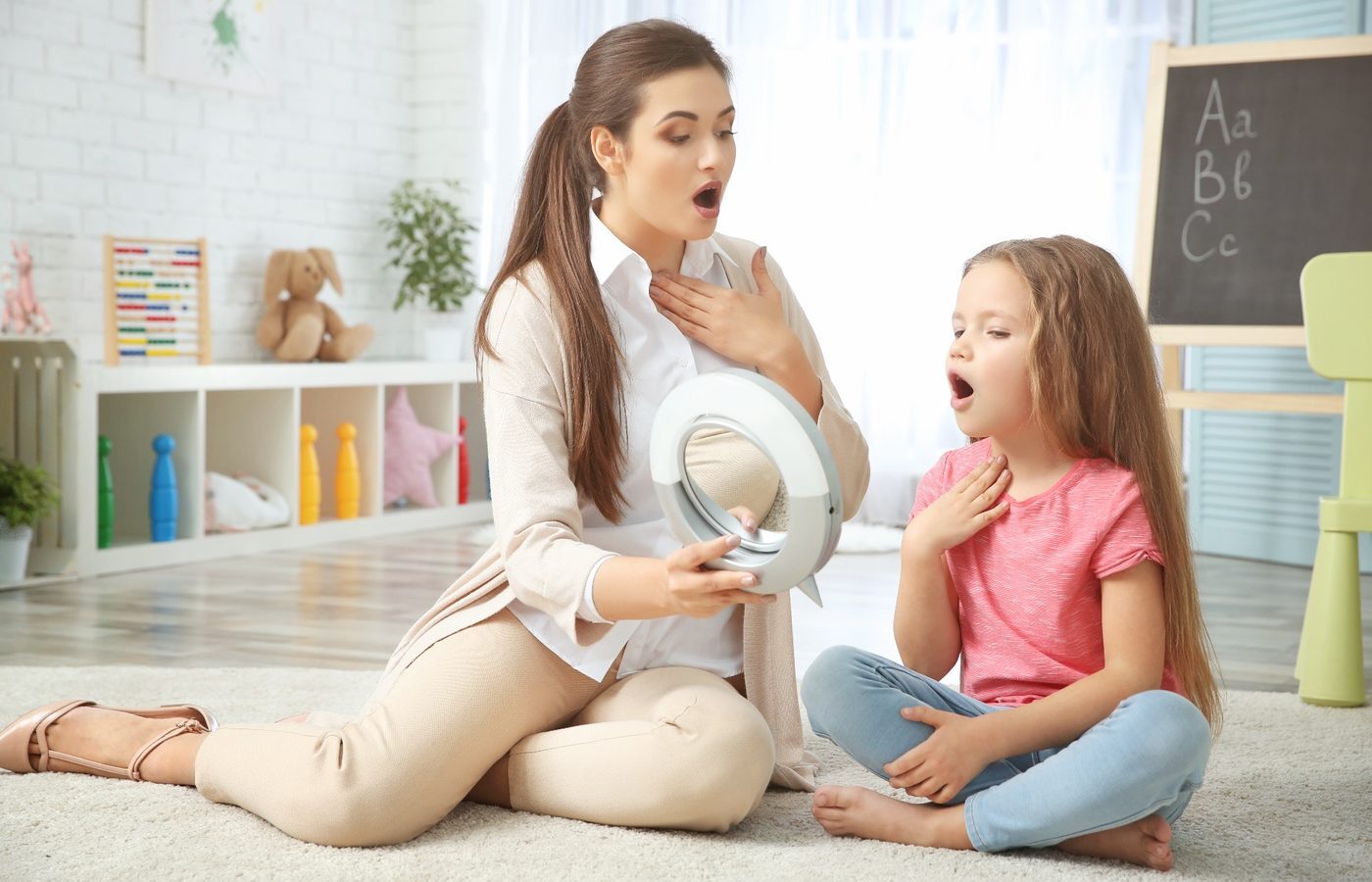 A speech-language pathologist holds a mirror while a young girl watches her own mouth shape a sound